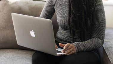 Person sitting on a couch using a silver Macbook; wearing a gray sweater and black pants.