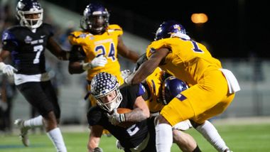 Football player in black uniform tackled by players in gold uniforms on a field at night.