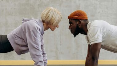 A woman and a man doing planks, facing each other. They are indoors, near a concrete wall.