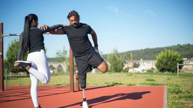 Woman and man stretching legs outside. Man has prosthetic leg, both touching support bar.