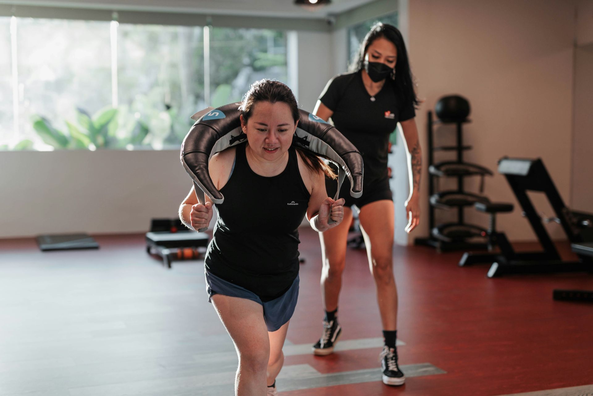 Woman doing a weighted carry exercise, guided by a masked trainer in a gym.