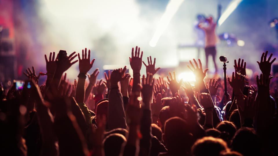 Audience with raised hands at a concert; stage with musicians in background, bright lights.
