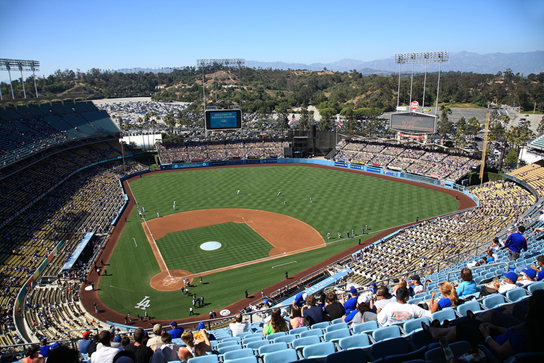 A wide view of Dodger Stadium filled with baseball fans. Green field, blue seats, sunny day.