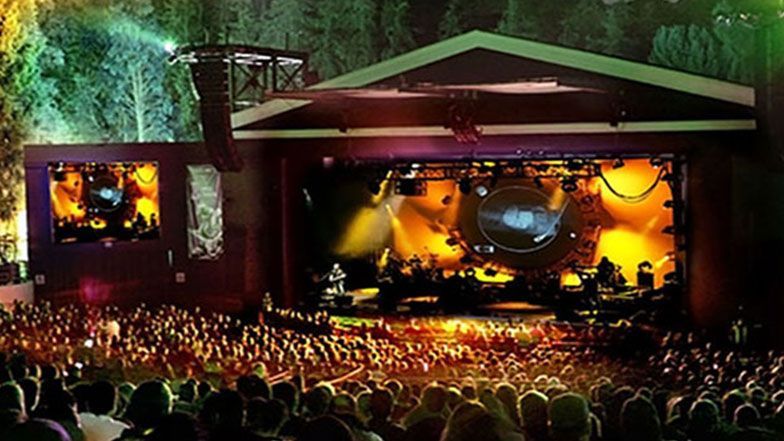 Concert at outdoor amphitheater: Band on stage with light show, large crowd in front.