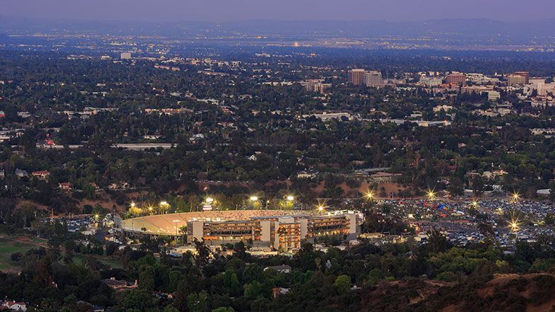Night view of a stadium with bright lights in a city, nestled in a valley of trees. Buildings and cityscape in the distance.