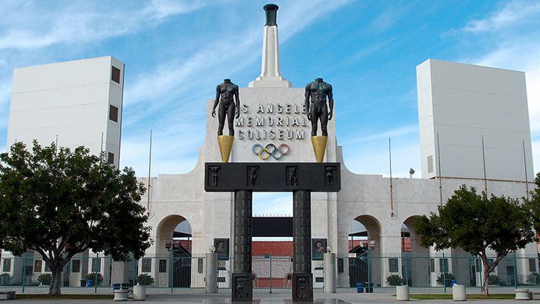 Los Angeles Memorial Coliseum entrance with statues, Olympic rings, and monument under a blue sky.