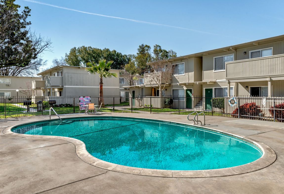 Apartment complex with a swimming pool. Tan buildings, green pool, blue sky.