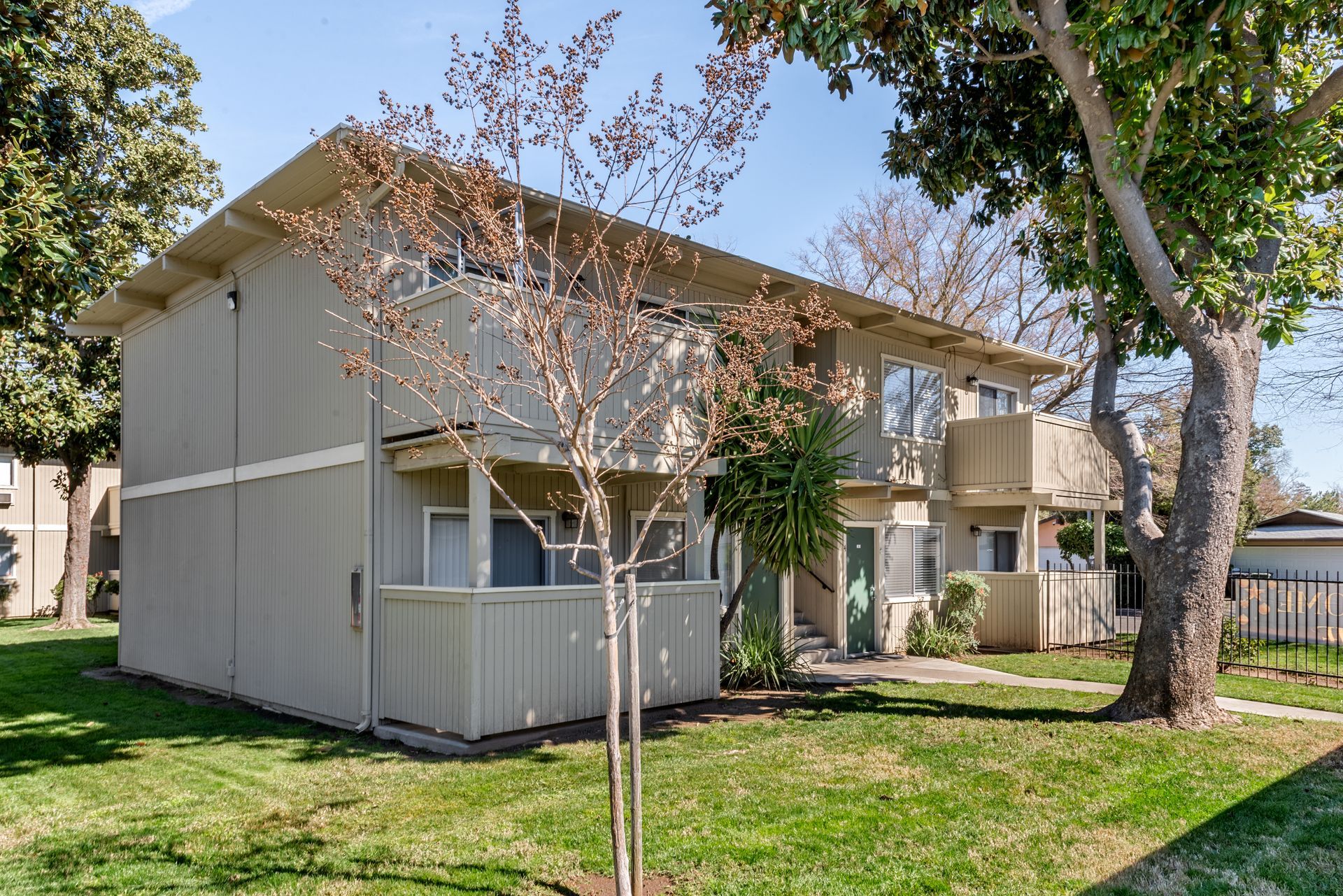 Two-story apartment building with tan siding, green lawn, and trees on a sunny day.
