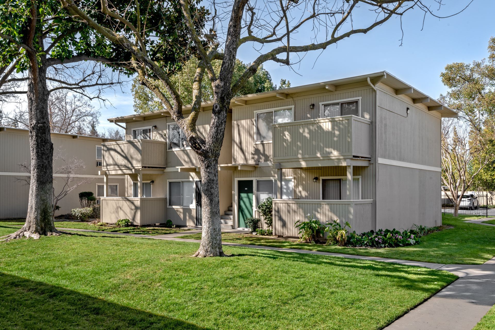 Two-story apartment building with beige siding, balconies, green lawn, and trees.