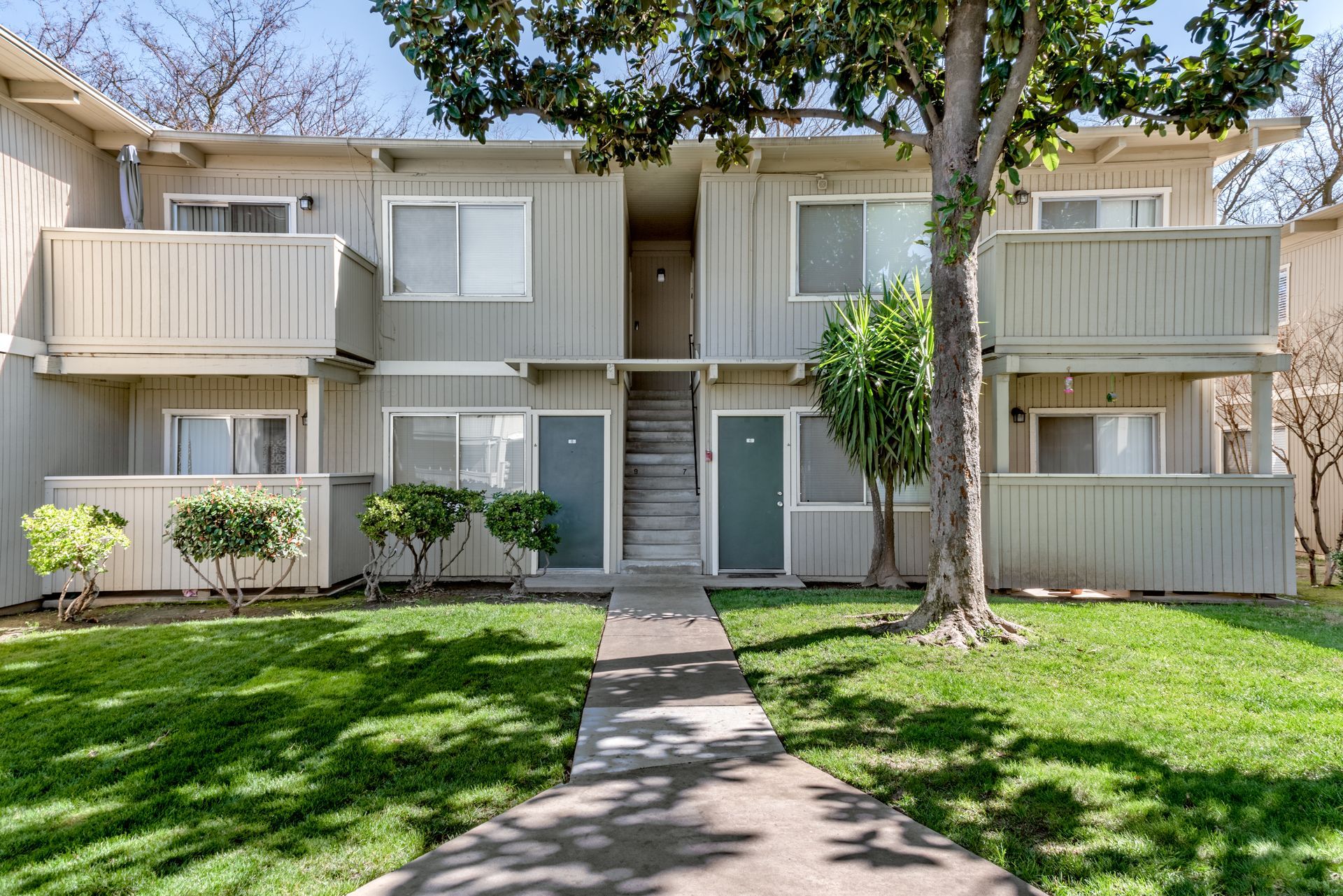 Apartment complex exterior with two-story units. Light beige siding, green grass, and a tree in front.