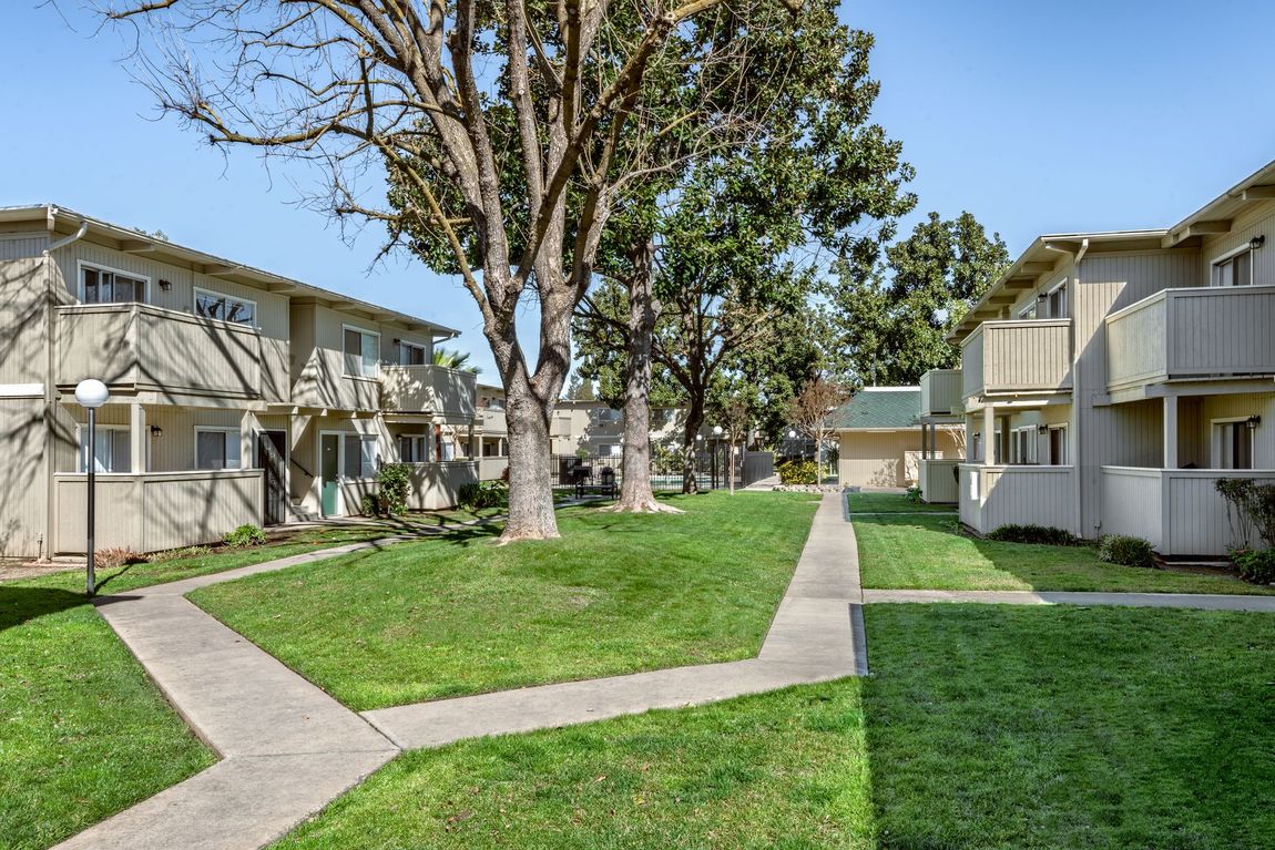 Apartment complex with two-story beige buildings, green lawns, and walkways lined with trees.
