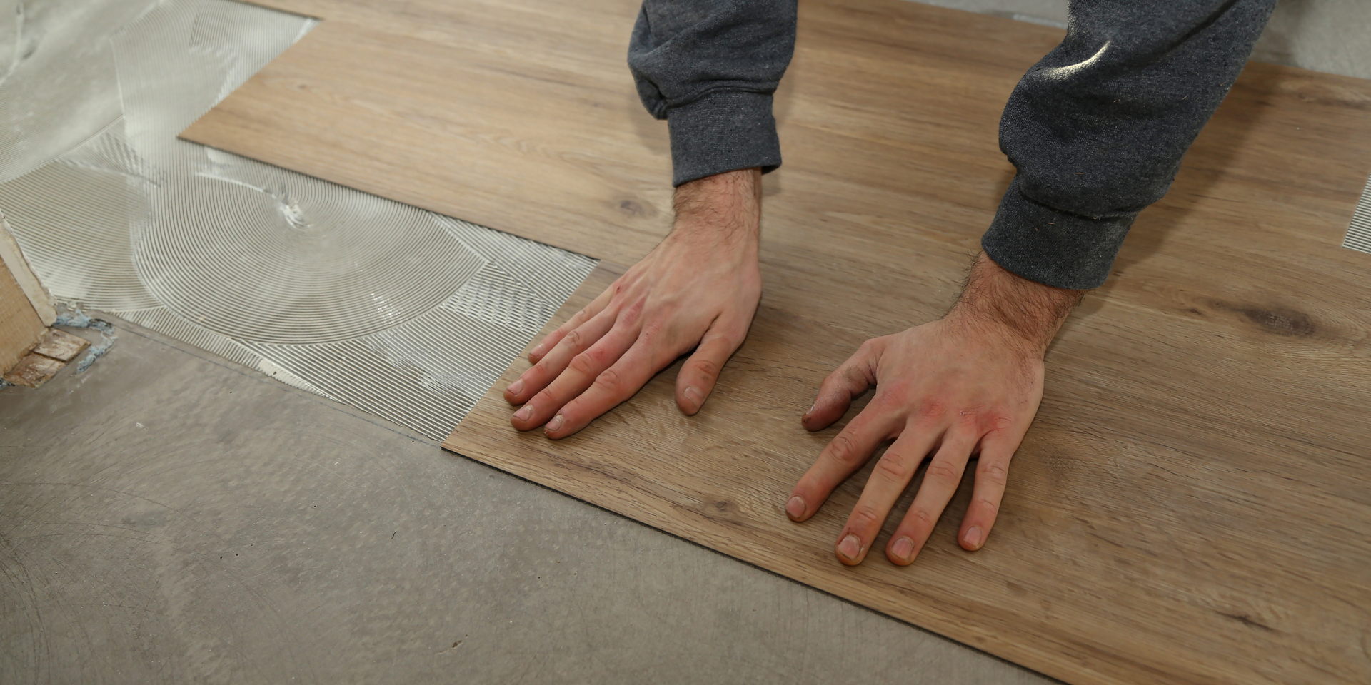 Close-up of hands installing wood-look vinyl flooring over adhesive spread on a concrete floor.