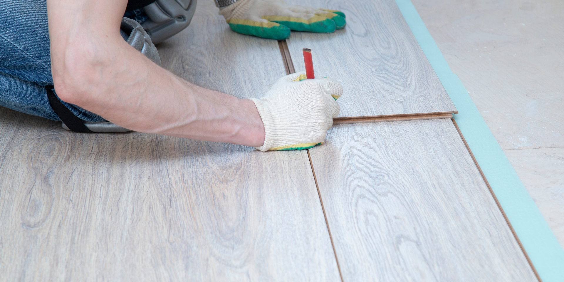 A person wearing gloves uses a tool to install light-colored wood laminate flooring planks on a floor.