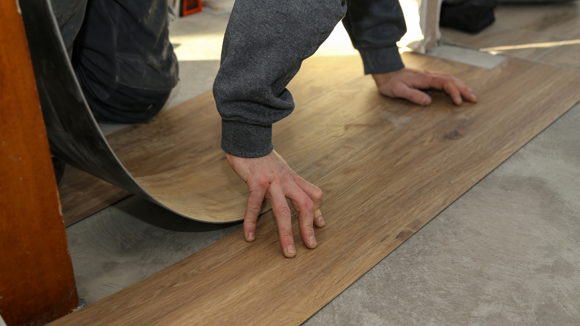 Person installing wood-look flooring, pressing the plank onto the floor. Gray sweater, concrete floor, indoor setting.