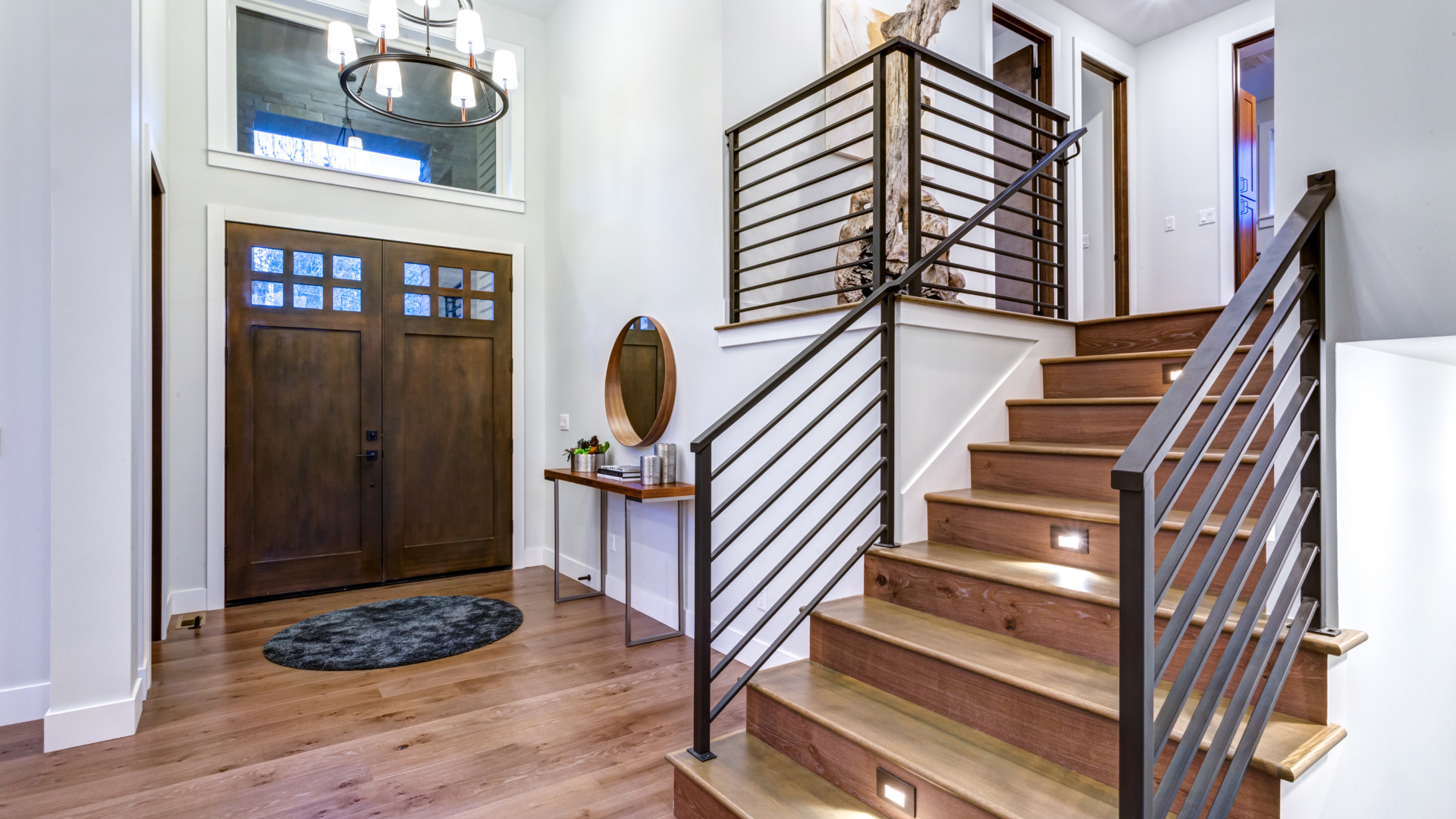 A modern entry hall with a wooden double door, a staircase with a horizontal metal railing, and a console table.