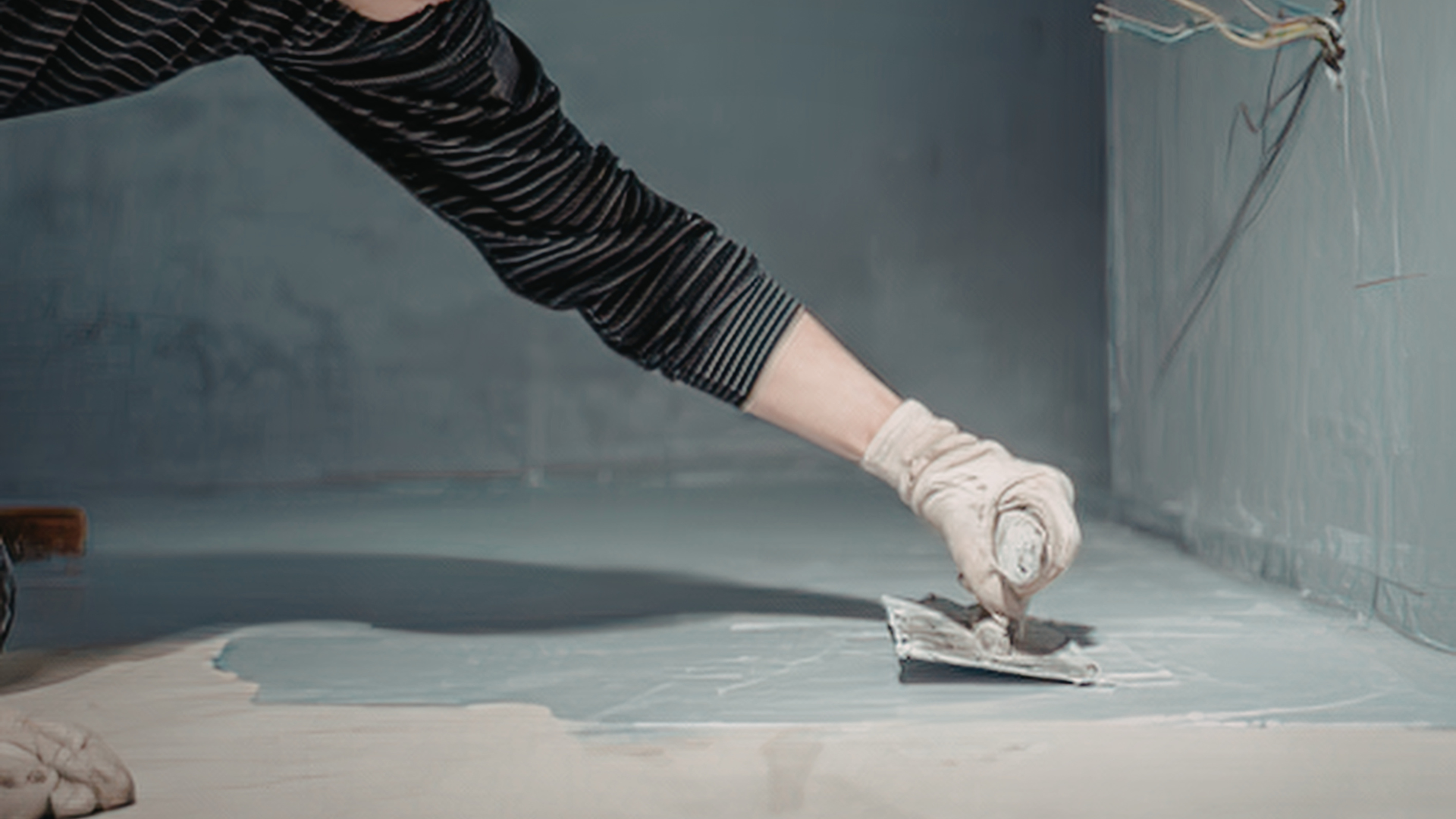 Person wearing gloves smoothing paint on a floor with a trowel, in a blue-walled room.