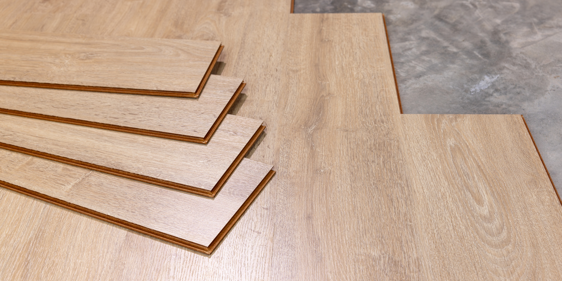 A stack of wood-look flooring planks sitting on a partially installed light-brown floor over a gray concrete subfloor.