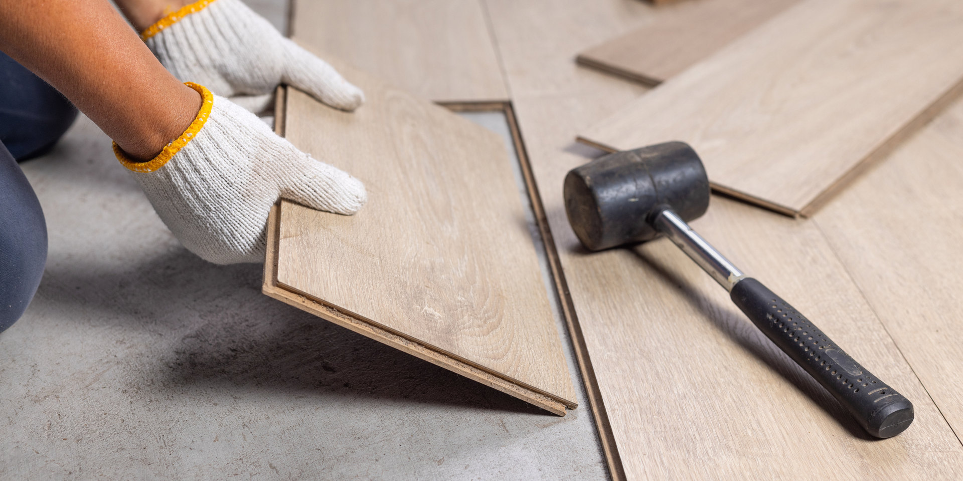 A person wearing gloves installing light-colored laminate flooring with a rubber mallet nearby.