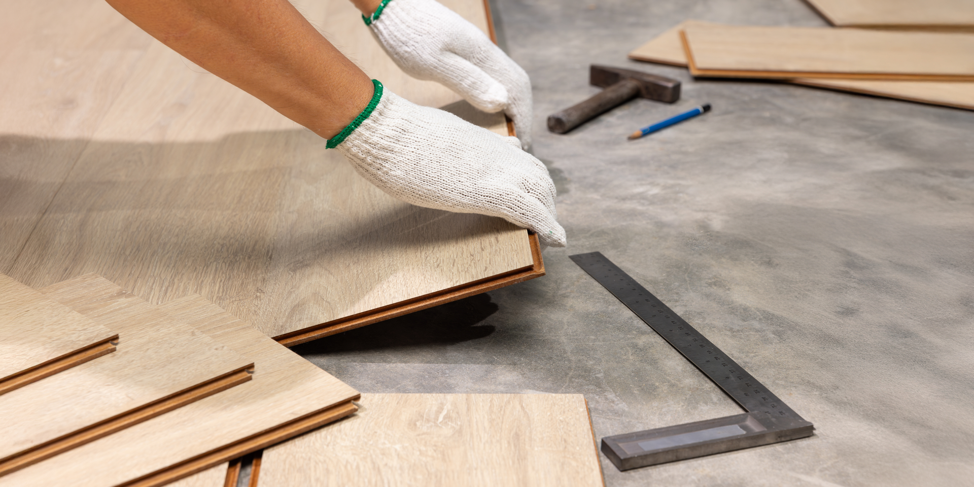 Hands wearing white gloves install light-colored laminate flooring, with a carpenter's square, pencil, and hammer nearby.