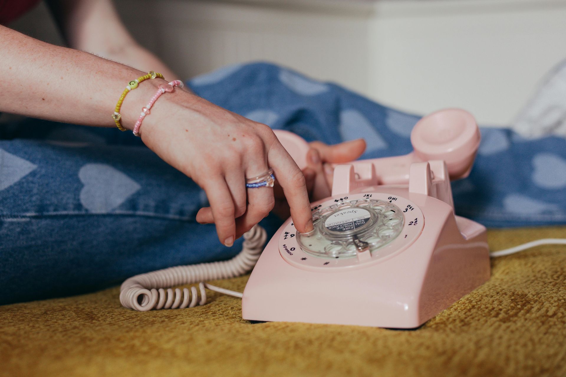 A person wearing beaded bracelets dials a number on a light pink vintage rotary phone while sitting on a textured surface.