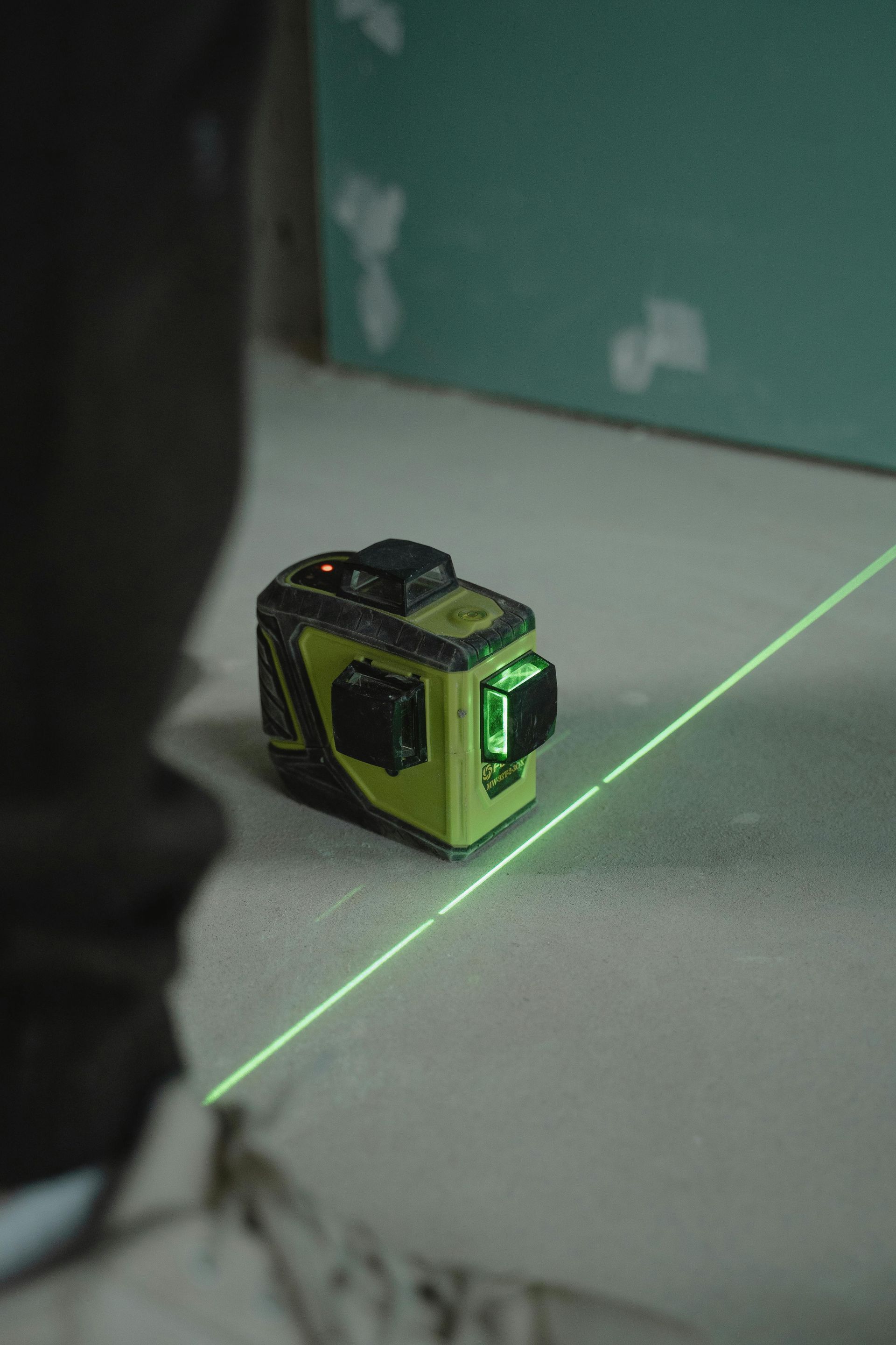 A green laser level projects a bright beam across a concrete floor near a person's feet and a green wall.