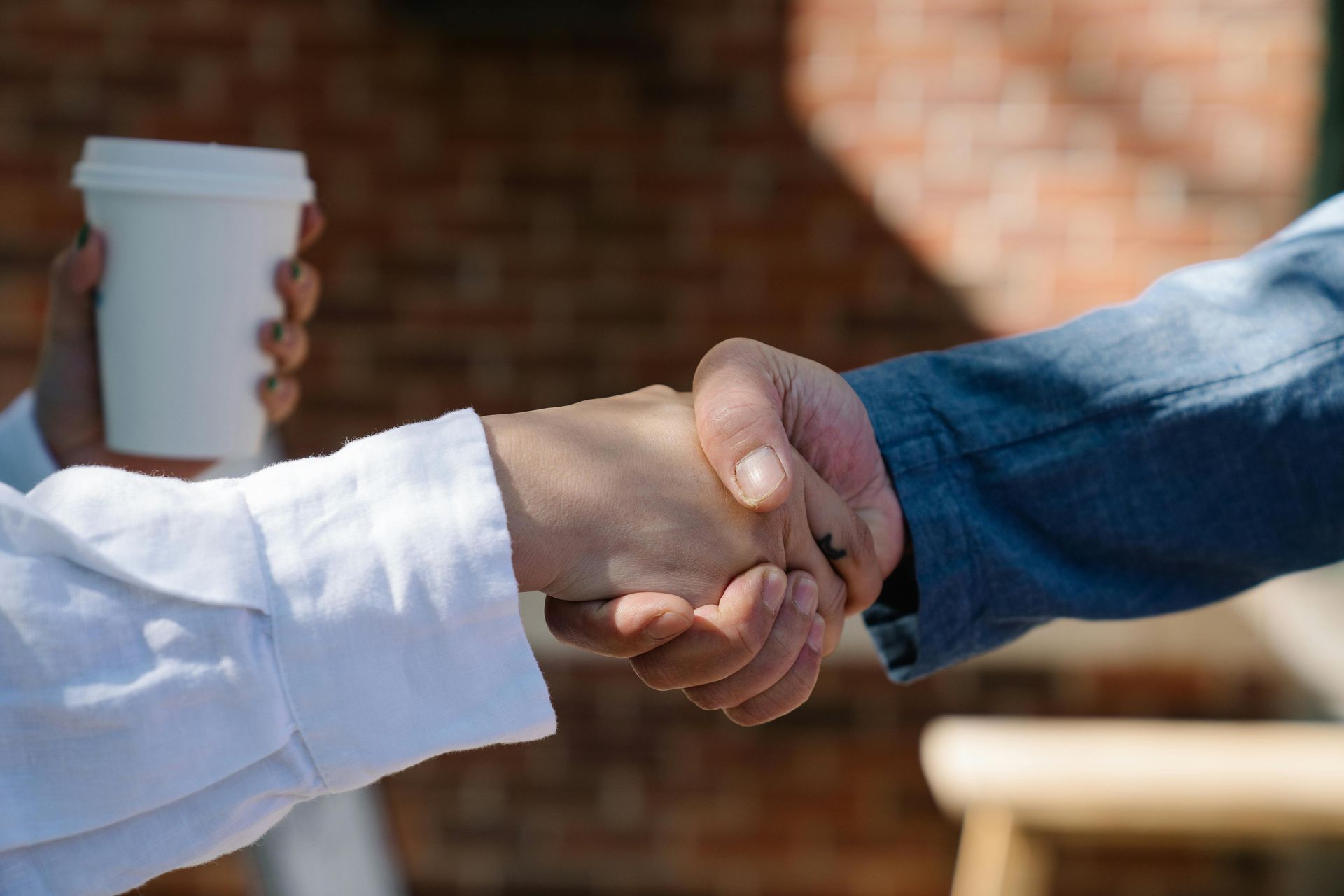 Two people shaking hands, with one person holding a white coffee cup, against a blurred brick background.