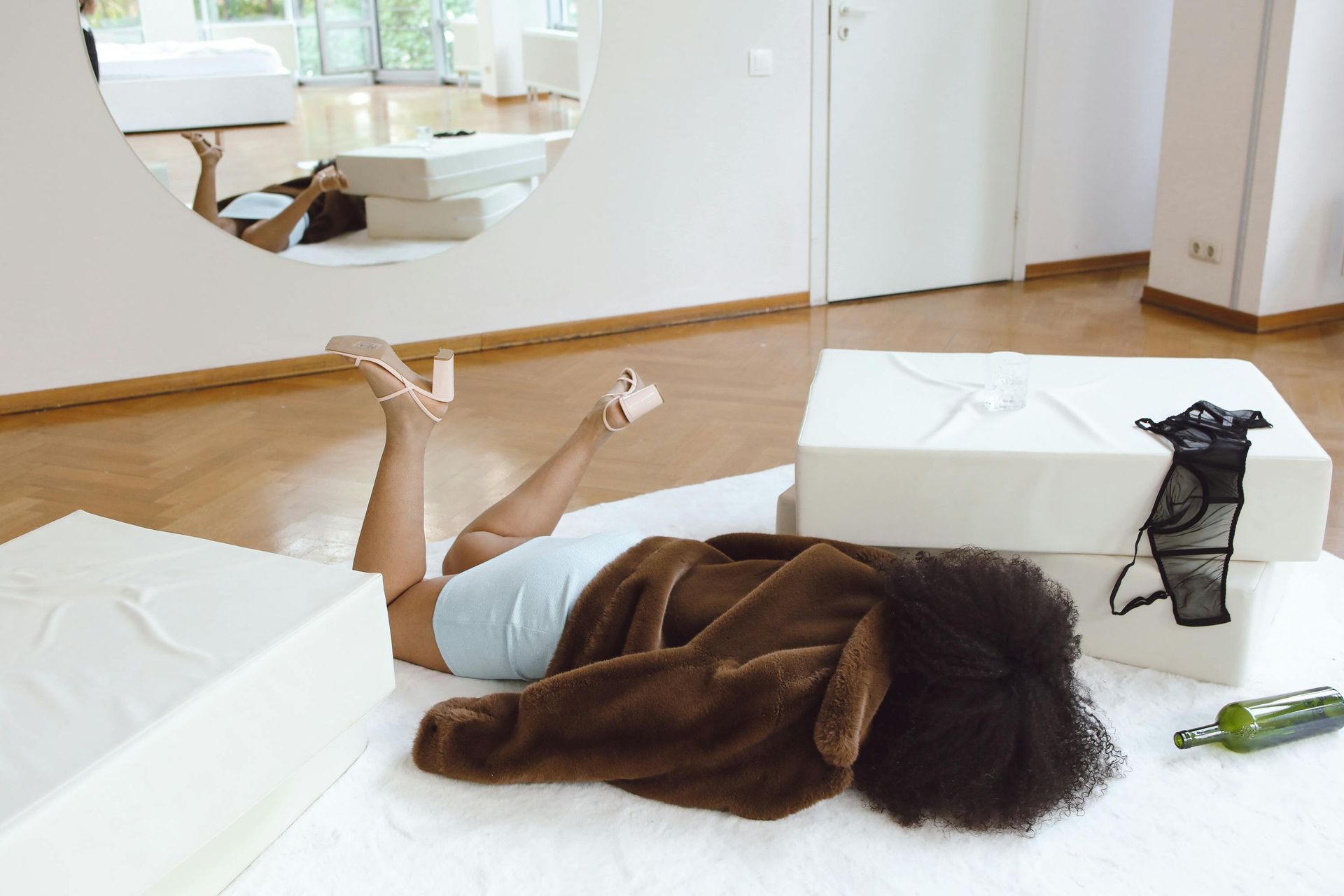 A person wearing a brown coat lies face-down on a white floor, with a black garment and a green bottle nearby.