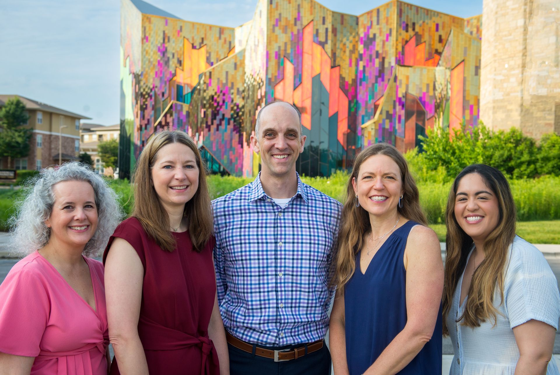 Five Pediatric Partners physicians stand together outdoors in front of a colorful geometric mural. They are smiling and dressed in professional, casual attire, with greenery and modern buildings in the background.