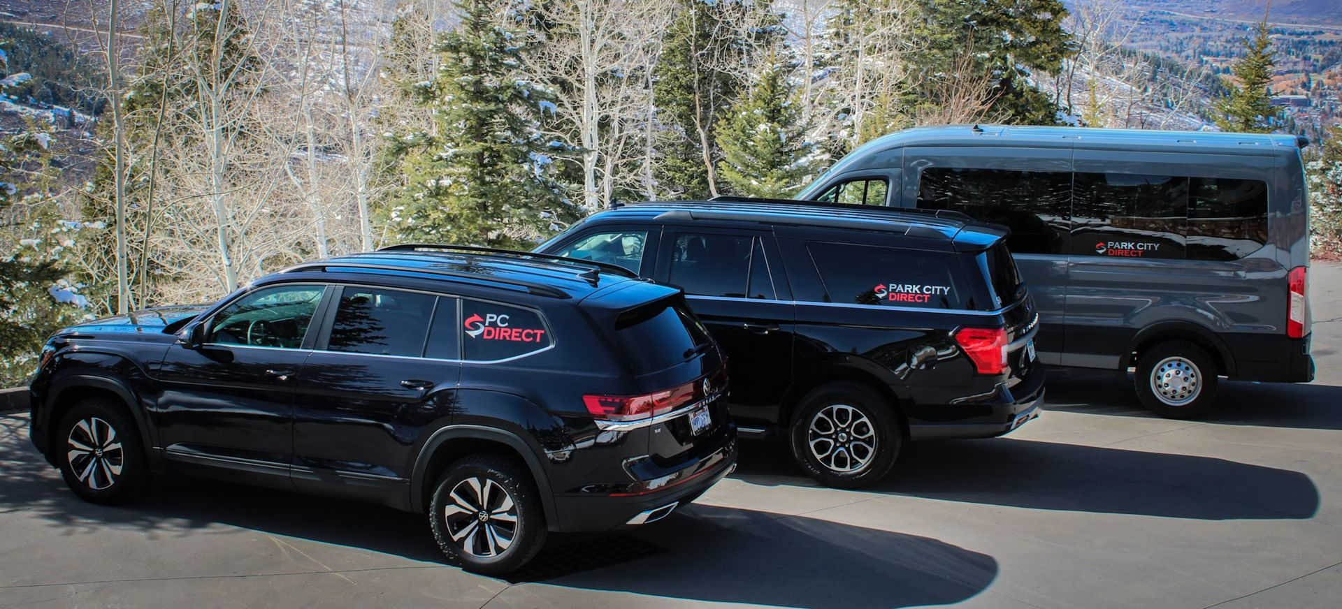 Three black vehicles parked on a paved road with a tree-lined hillside in the background.