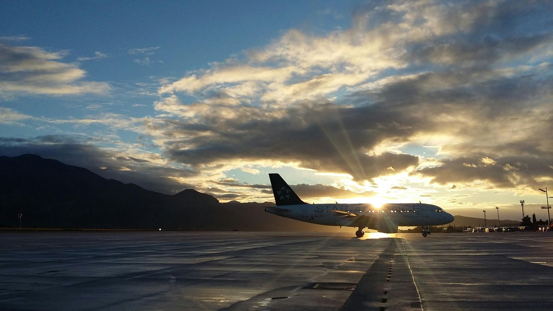 Airplane on runway with sunset, mountain in background. Silhouetted against bright sky.