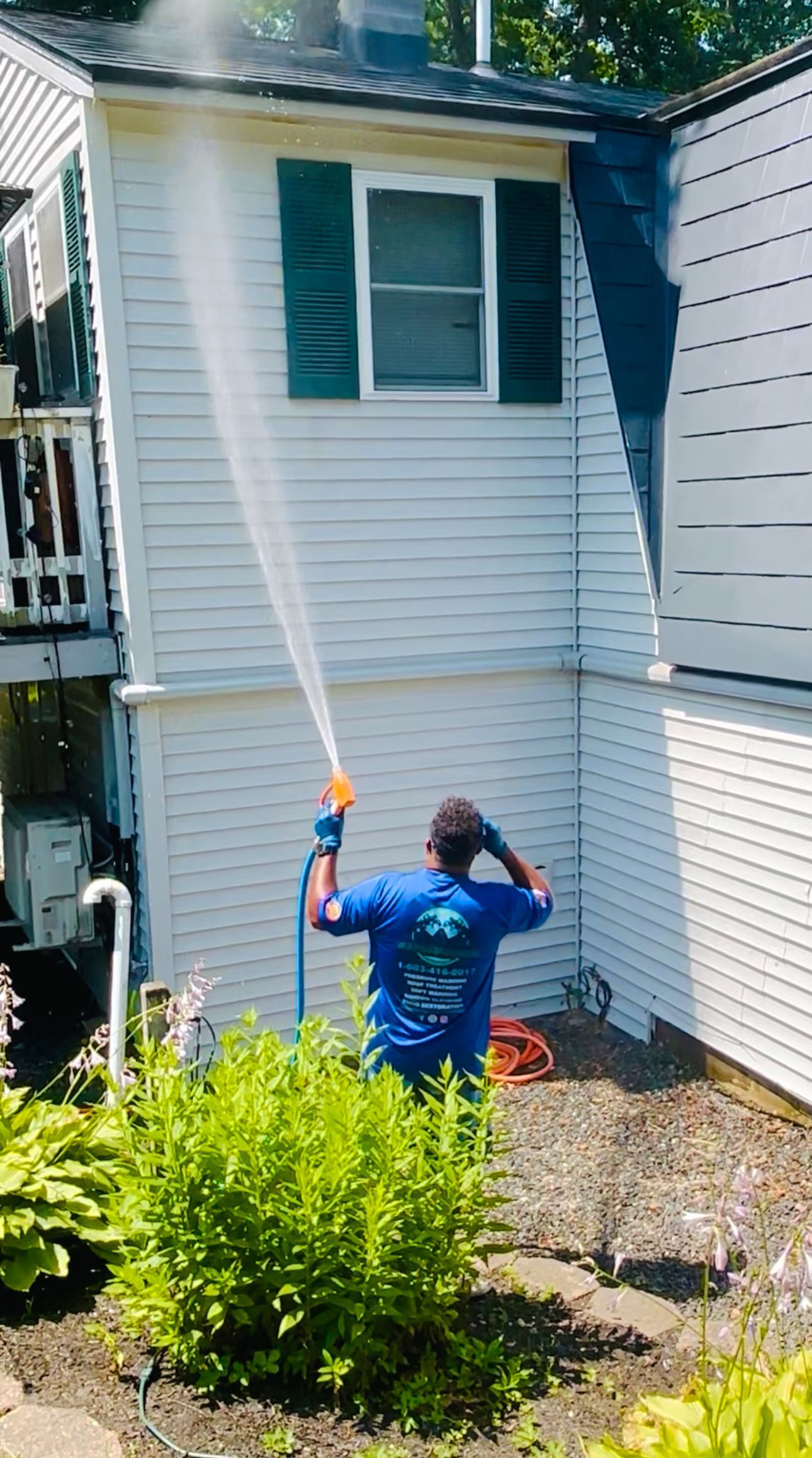 A person in a blue shirt uses a pressure washer to clean the white siding of a two-story house.