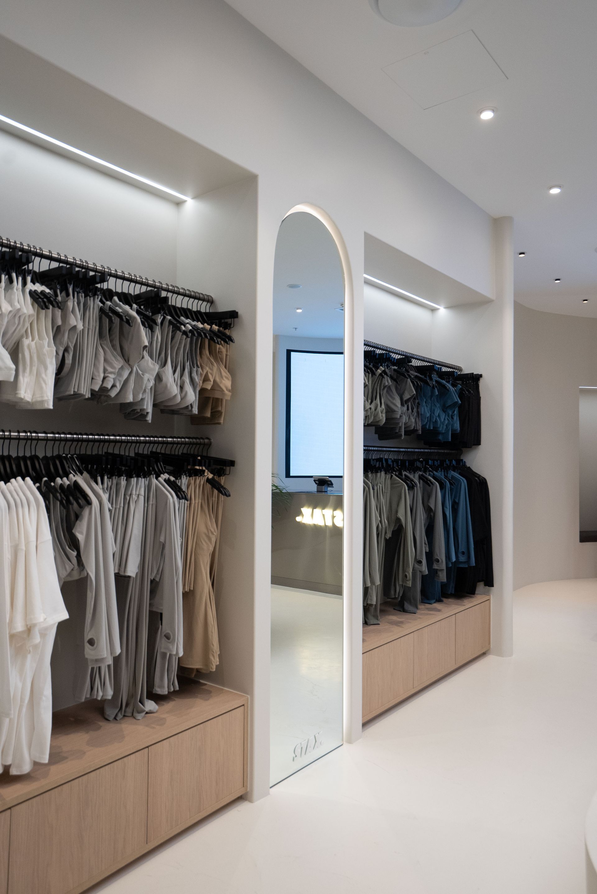 Clothing racks in a modern retail space with a large arched mirror reflecting a counter. Light wood and white interior.