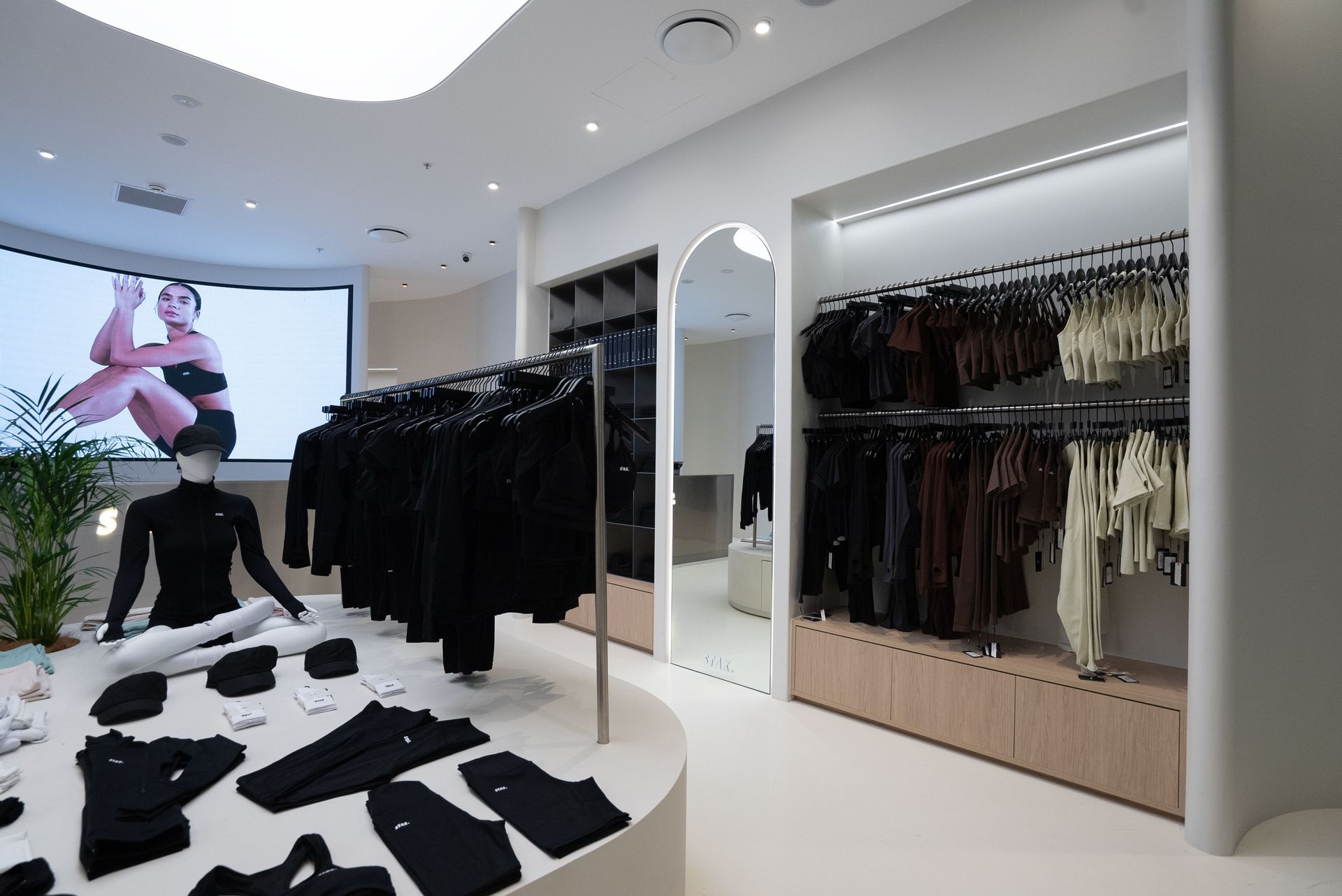 Clothing store interior with racks of black and brown apparel, display table, and large screen.