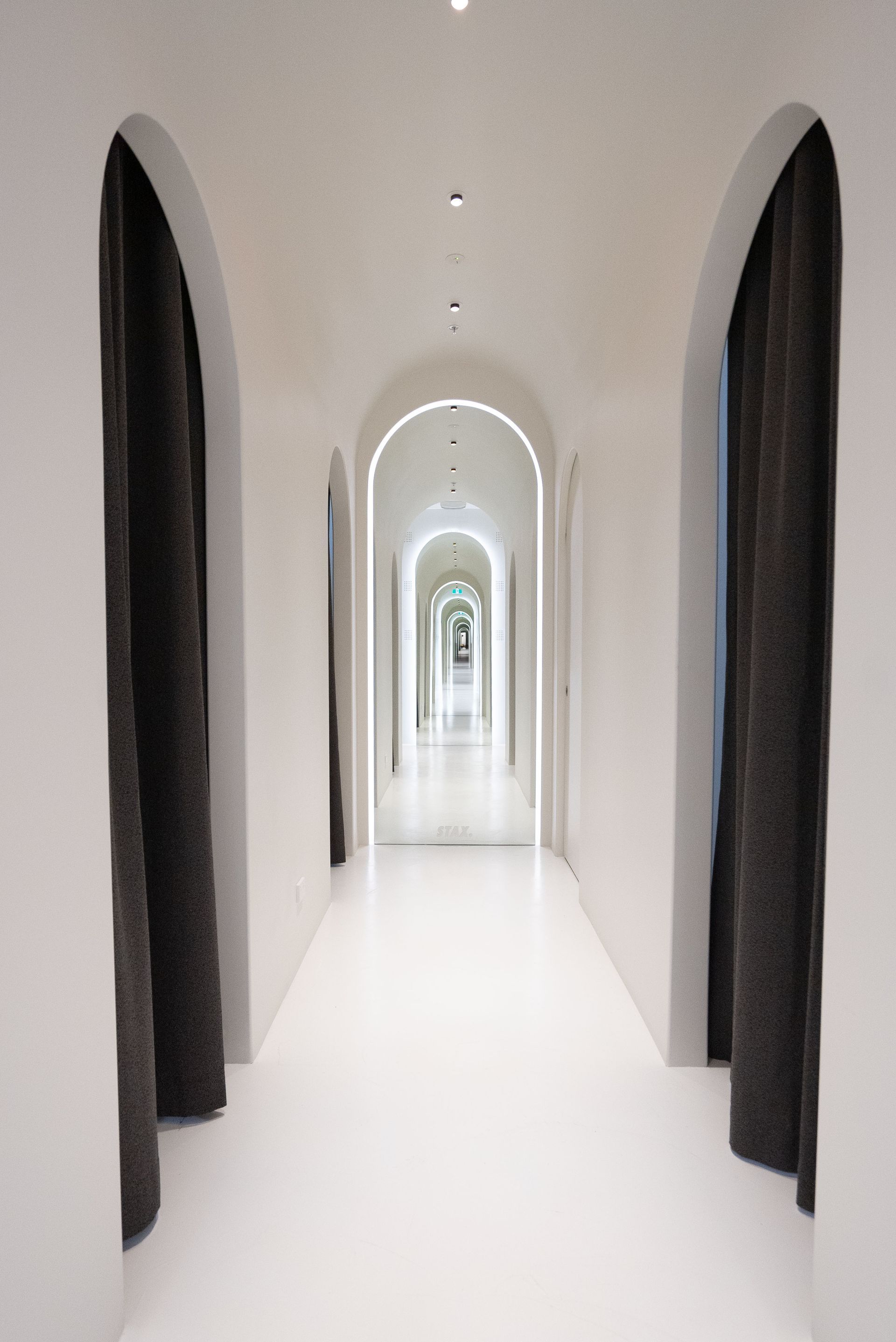 White hallway with arched doorways and dark curtains, leading to a bright, distant view.