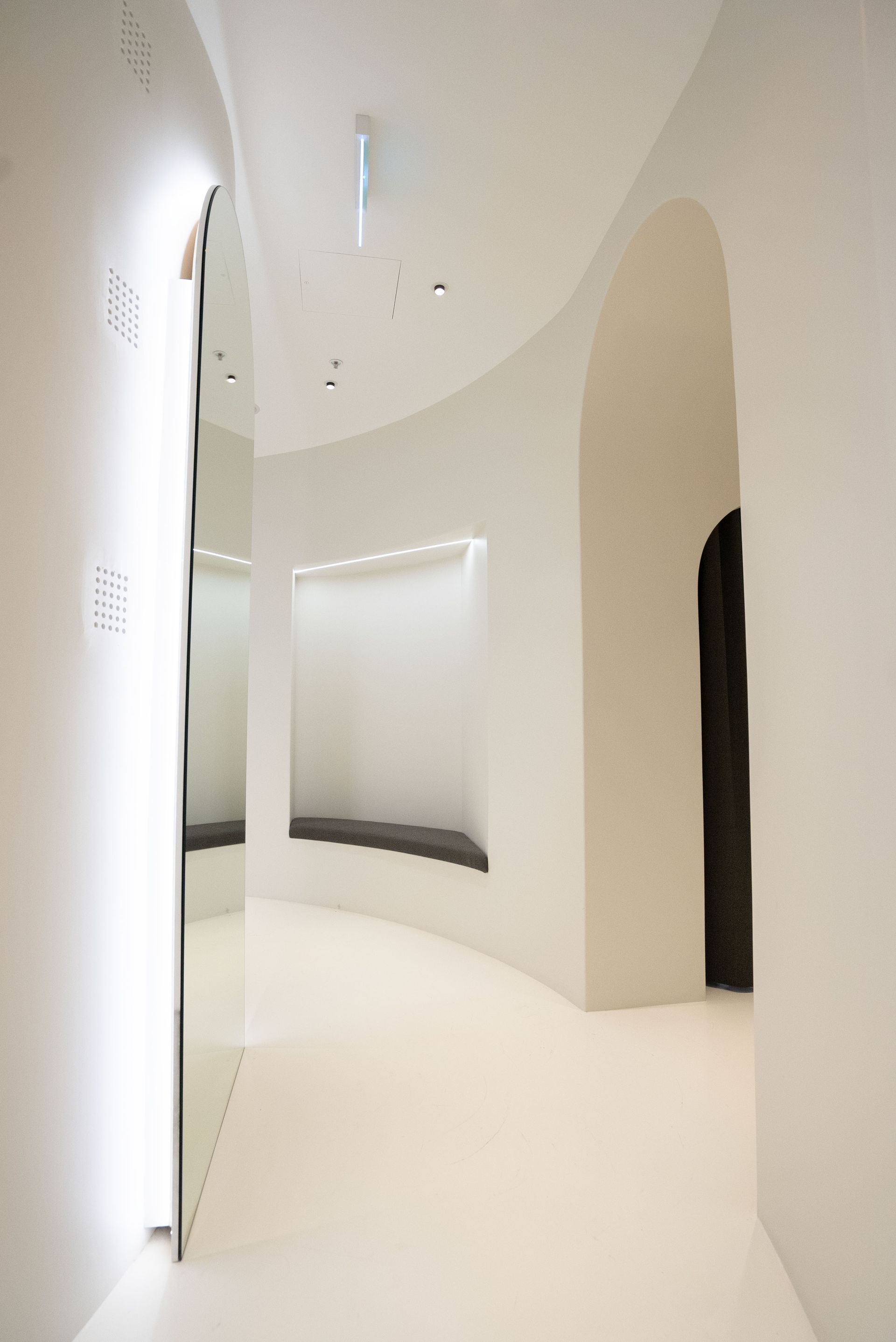 White curved hallway with a large mirror on the left and an arched doorway at the end.