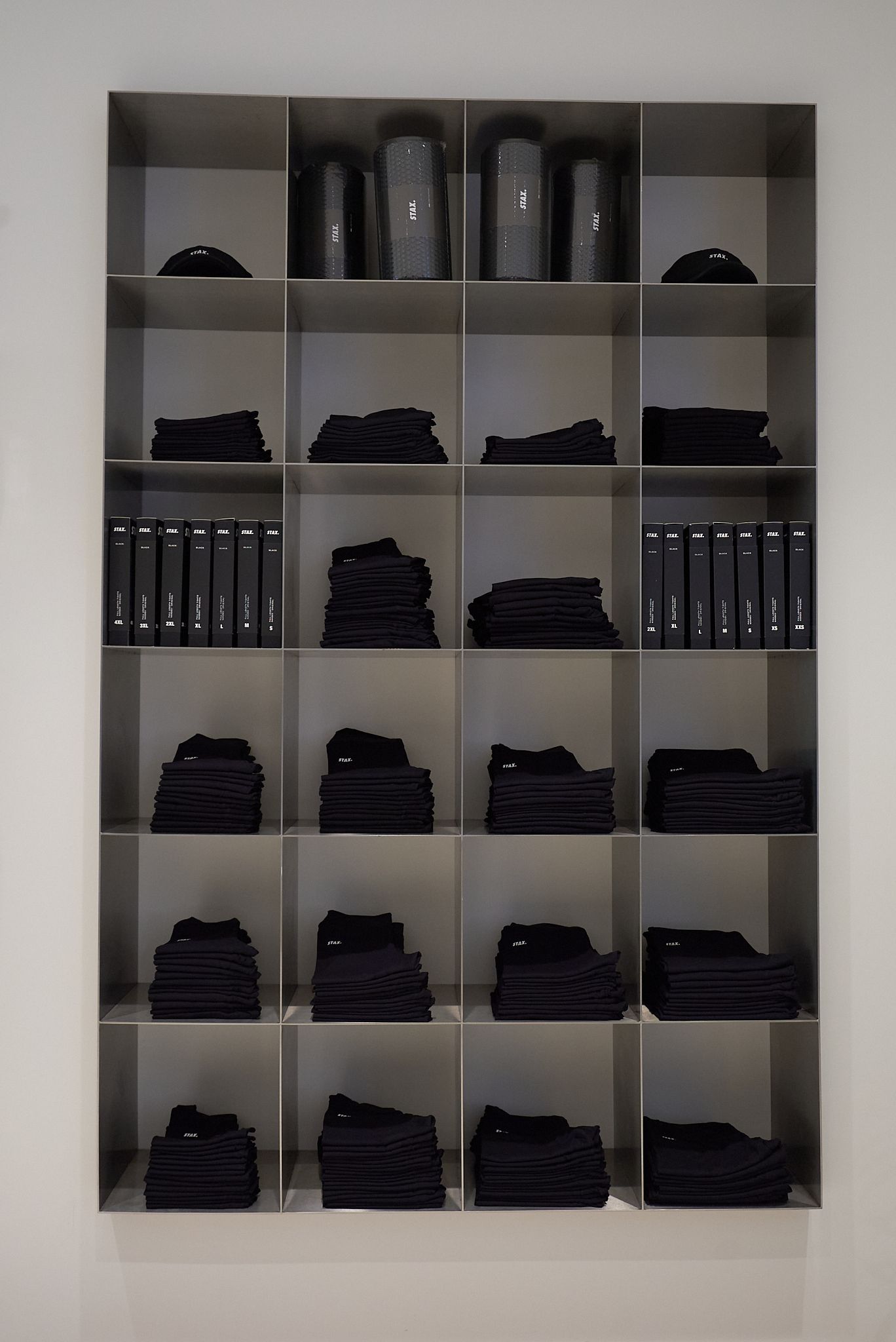 A metal shelving unit with 20 compartments, holding black objects: clothes, books, and cylindrical shapes.