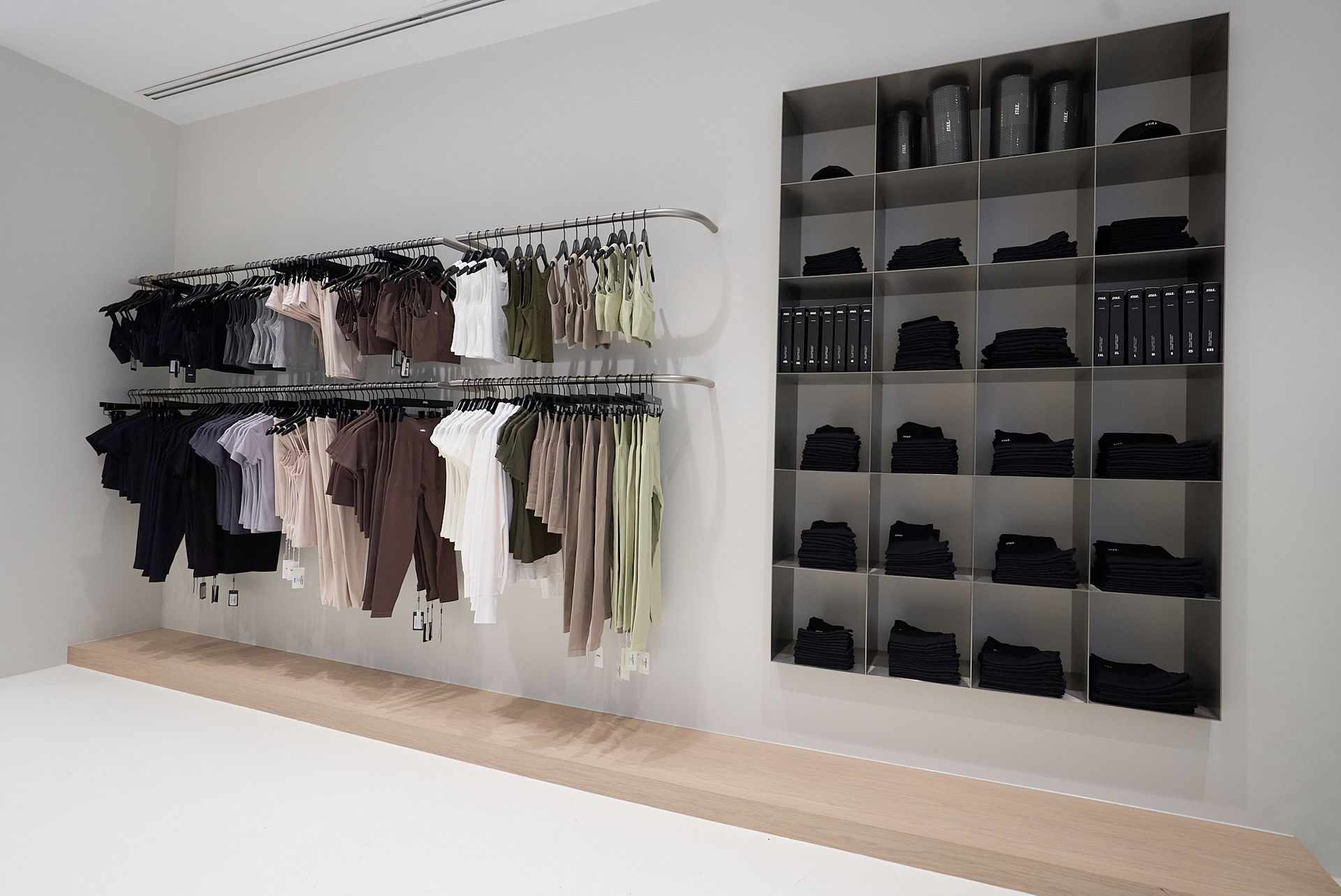 Clothing store display of neutral-toned apparel on racks and shelves against a light gray wall.