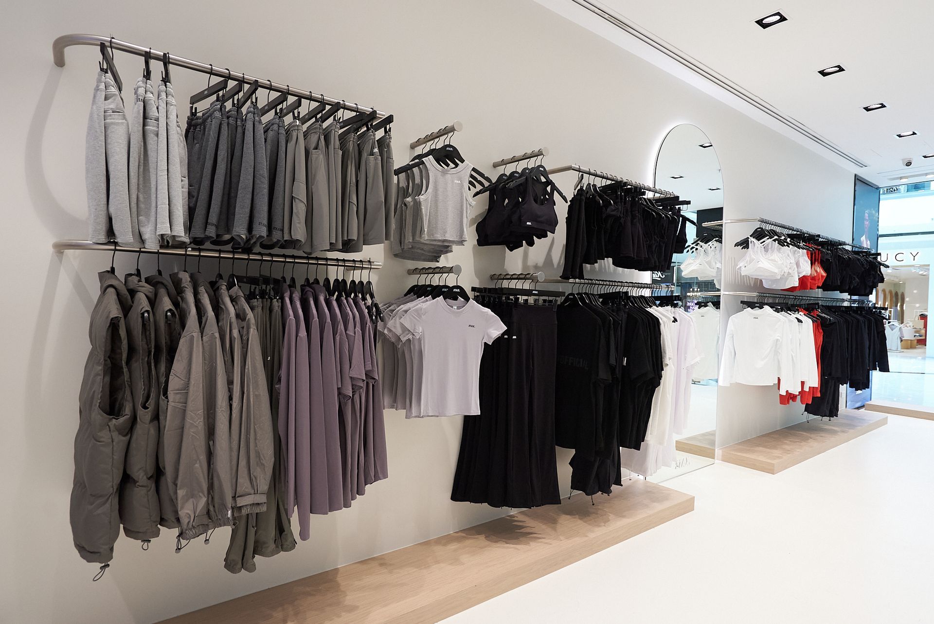Clothing store display with racks of gray, black, and white garments hanging against a white wall and floor.