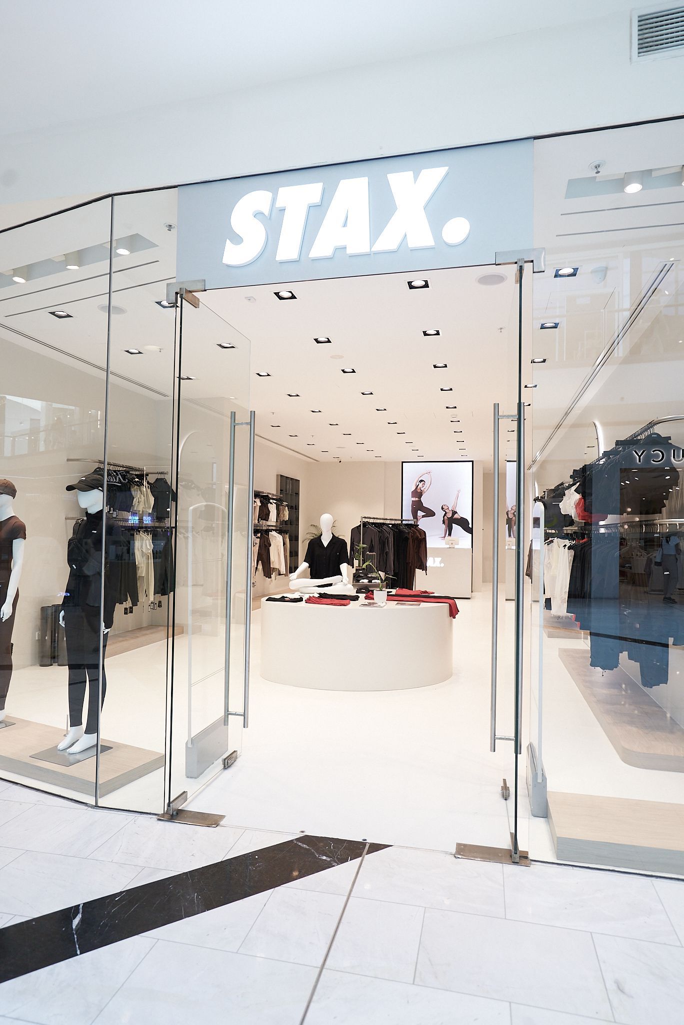 STAX. store front with glass doors, white walls, mannequins displaying clothing, and a central white counter.