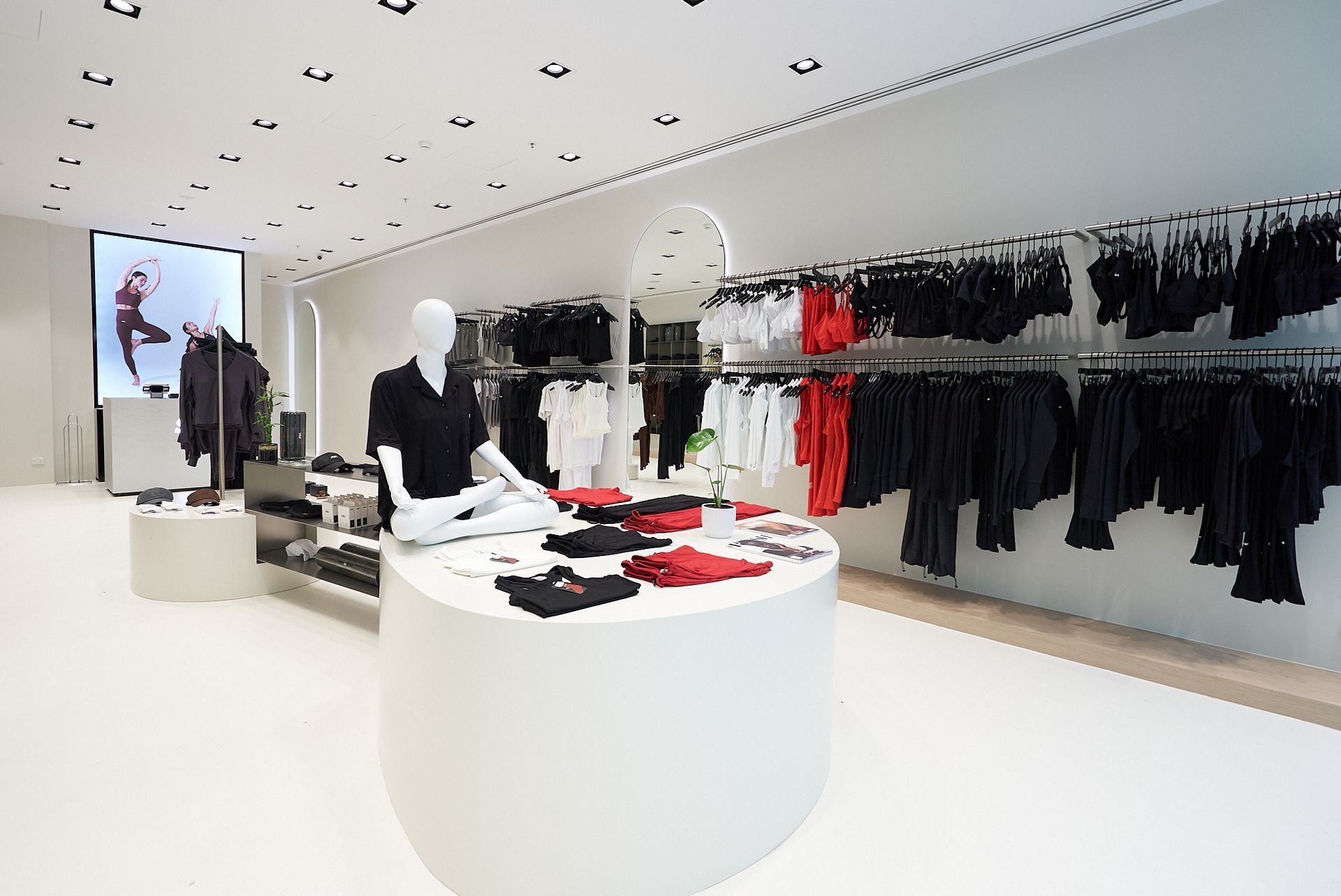 Inside a bright clothing store, mannequins and racks of black and red garments are displayed on white surfaces.