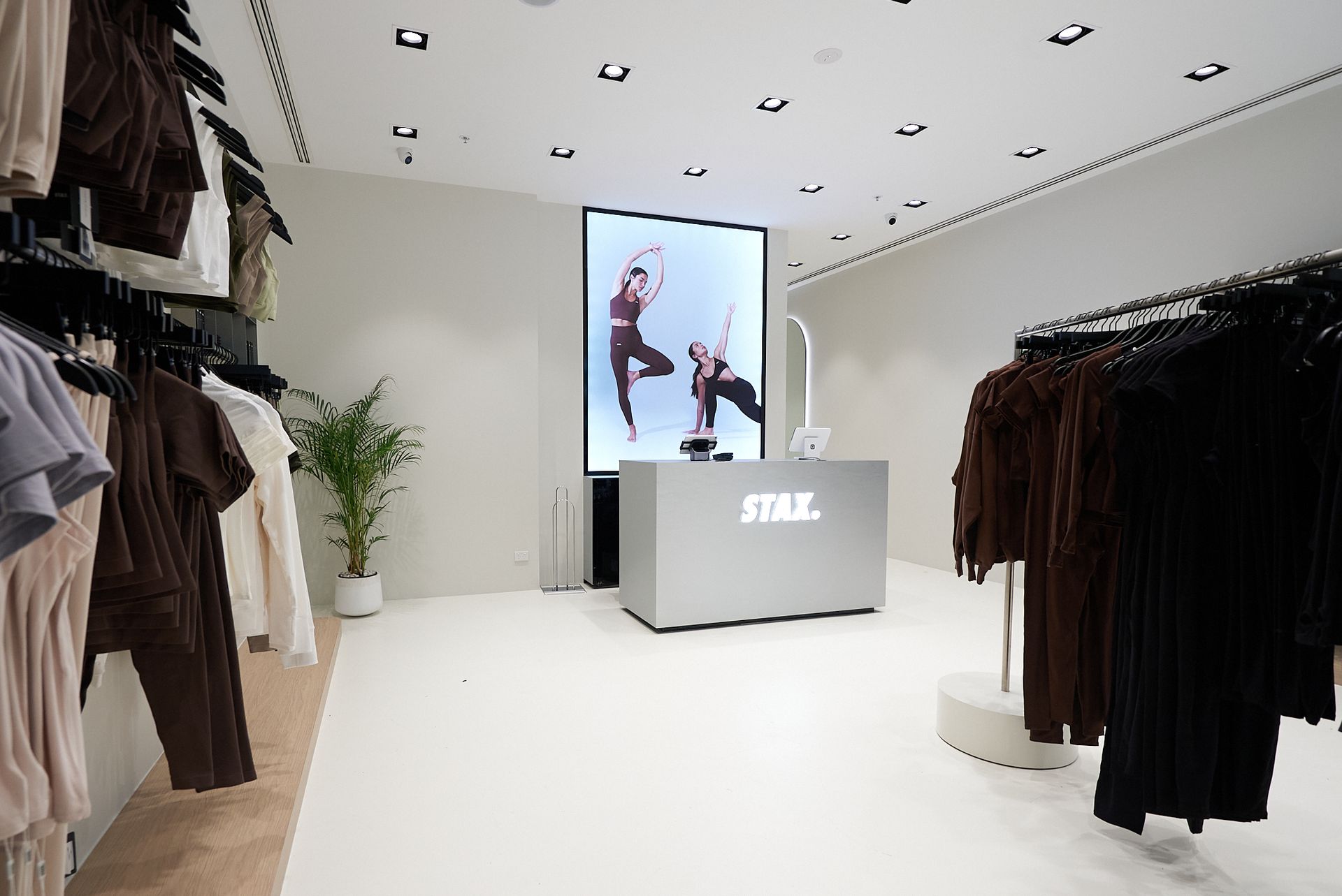 Clothing store interior with clothing racks on either side of the room. A central display screen shows dancers.