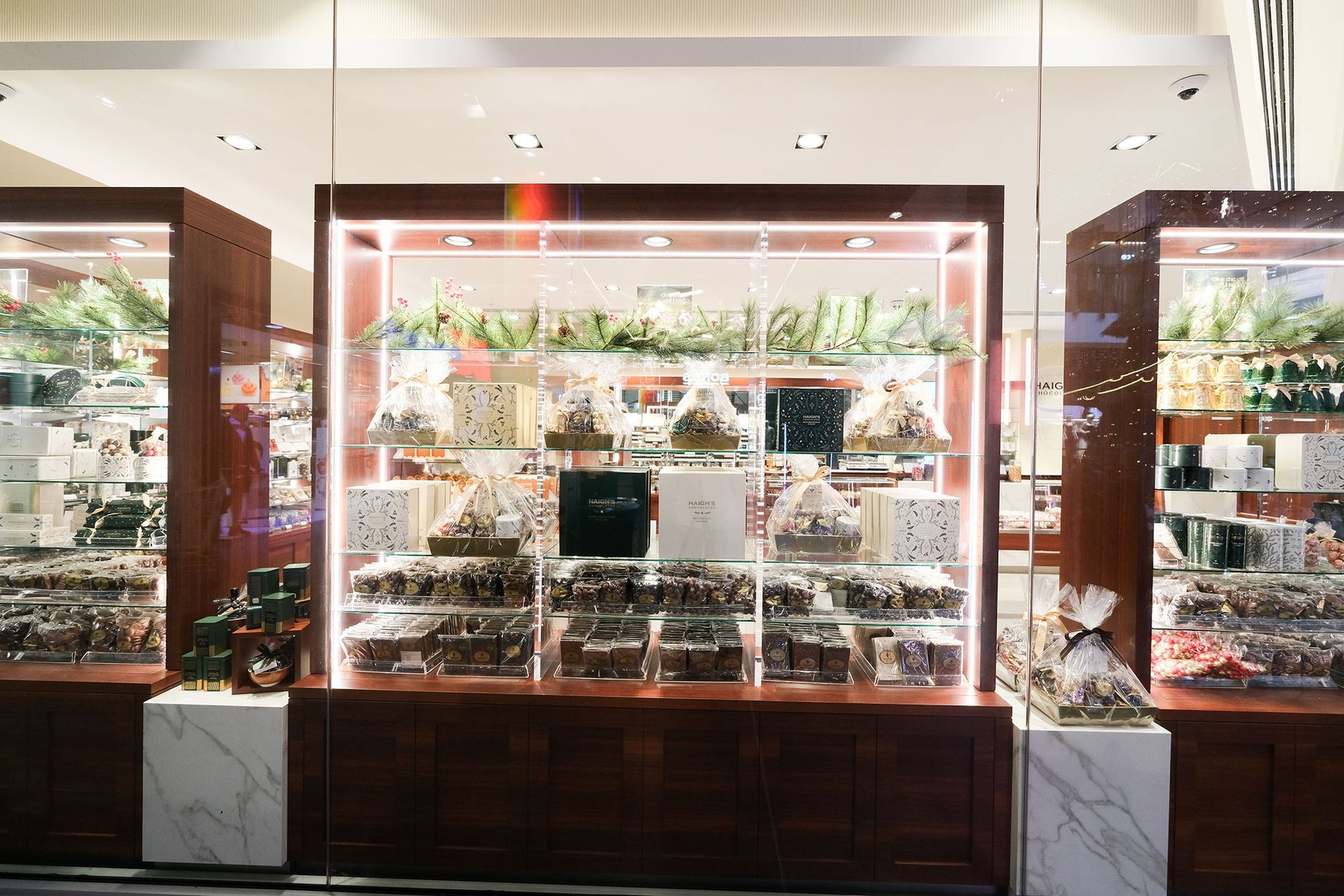 Chocolate shop display: Glass cases with various chocolates and gift packaging. Brown and white tones with bright lighting.