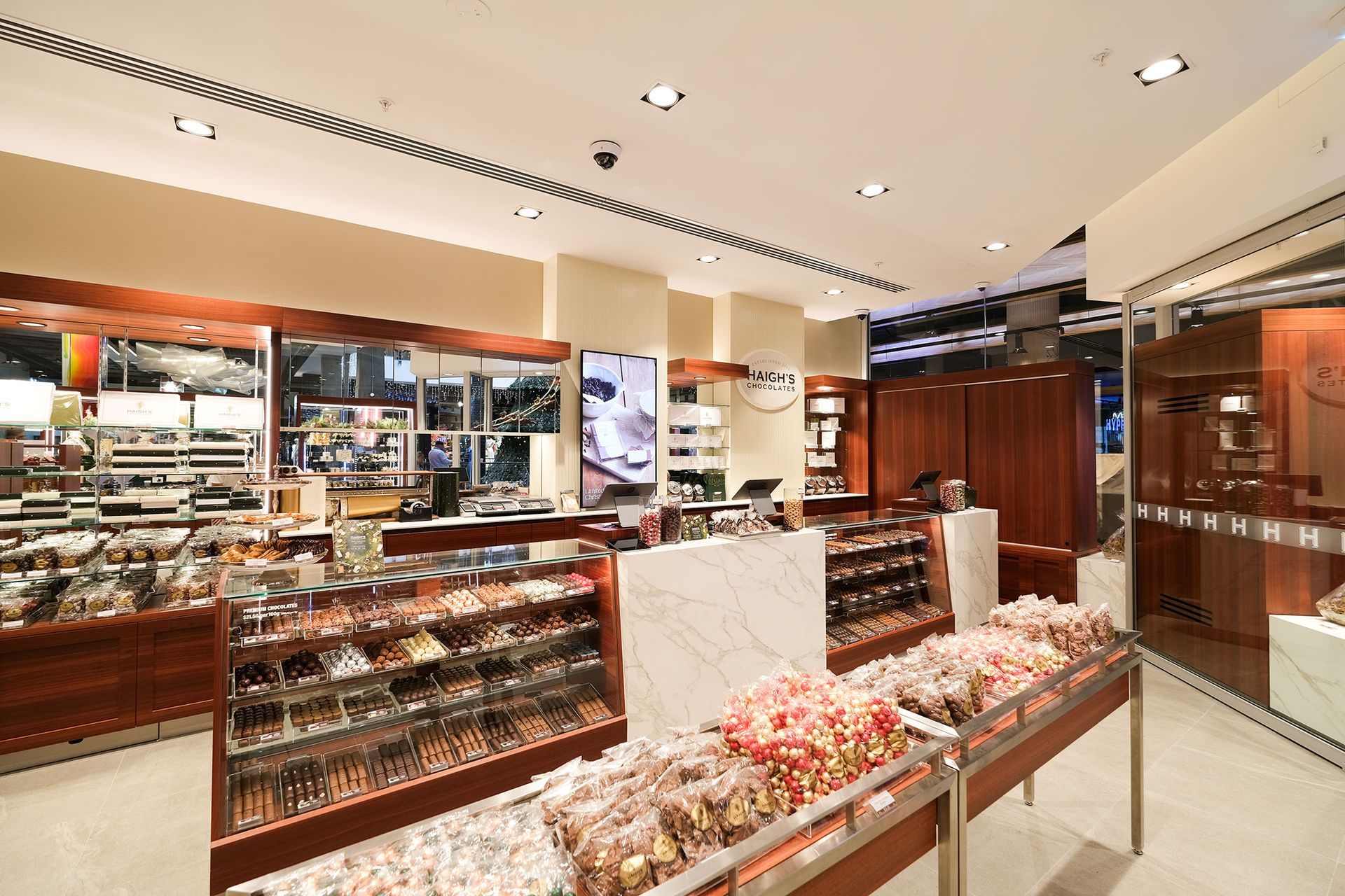 Inside a candy store: display cases filled with sweets, brown wooden shelves, marble counter, overhead lighting.