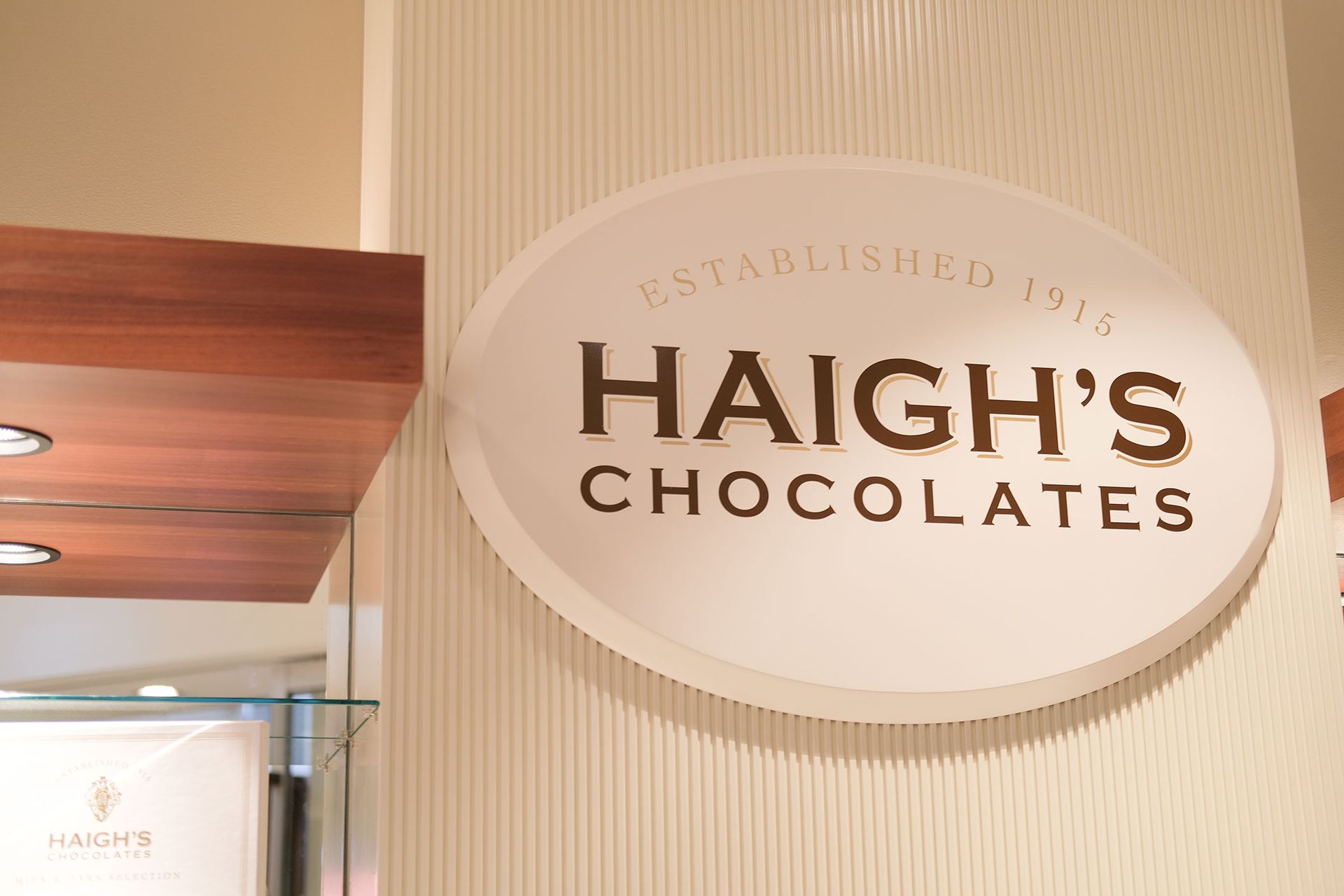 Oval sign for Haigh's Chocolates, established 1915, in gold lettering on a white background, mounted on a ridged wall.