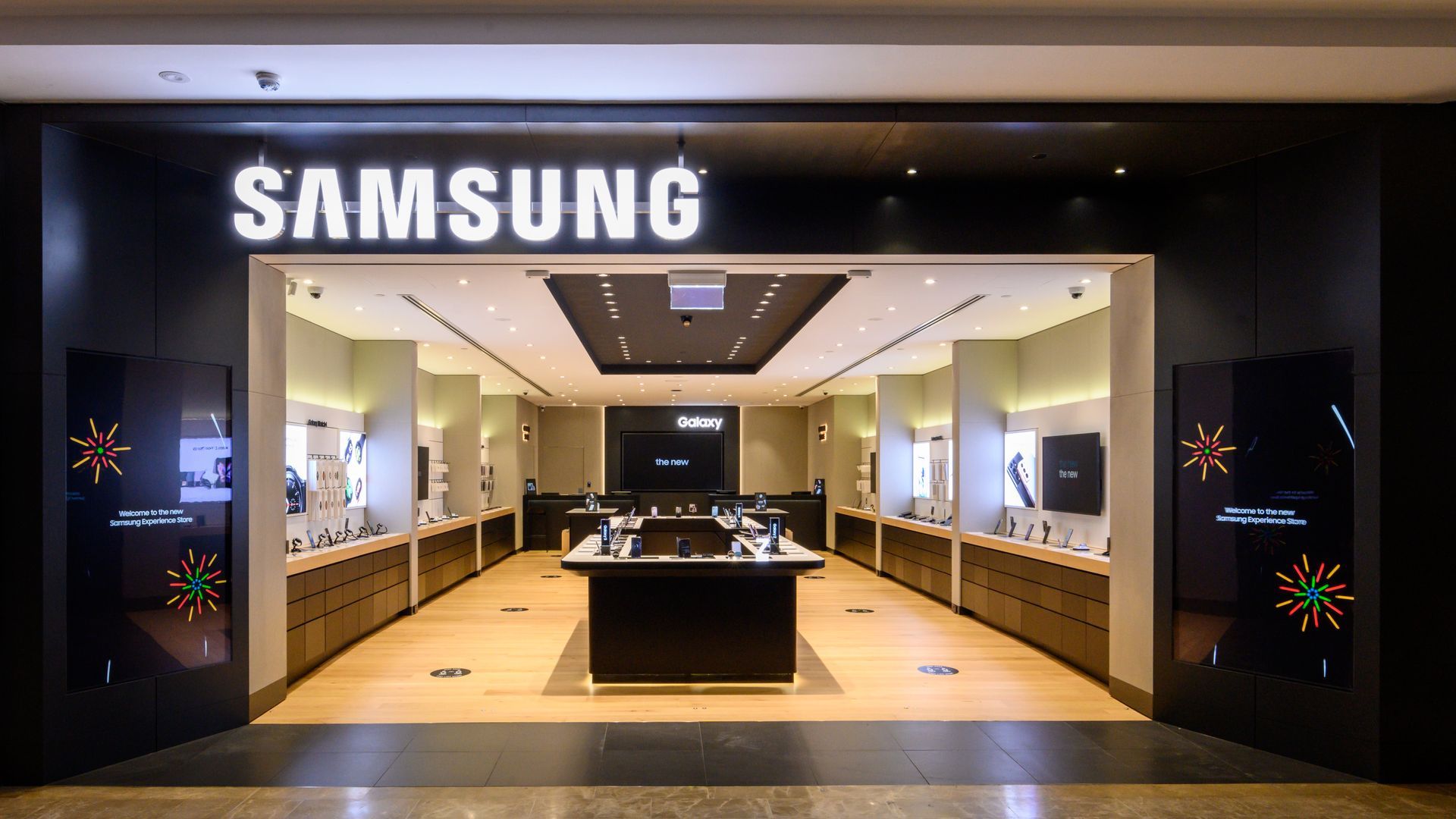 Entrance of a Samsung store, black facade with illuminated white logo, glass doors, interior displays.