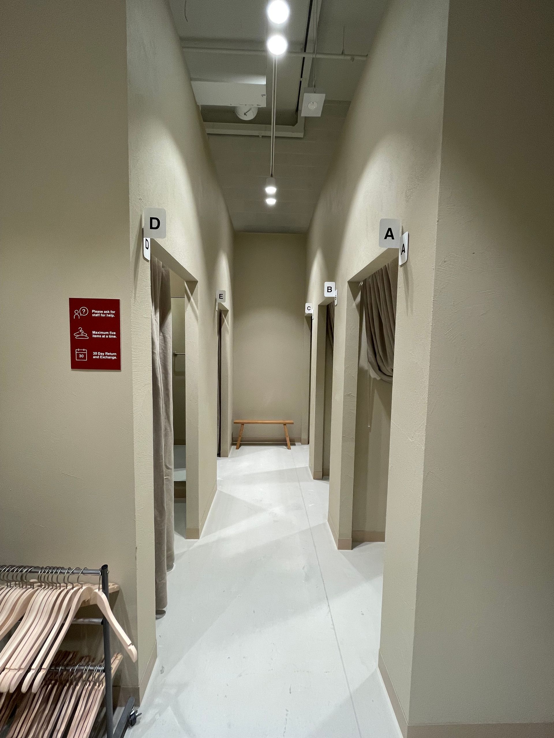 Narrow retail dressing room hallway, beige walls, white floor, curtained stalls, overhead lighting.