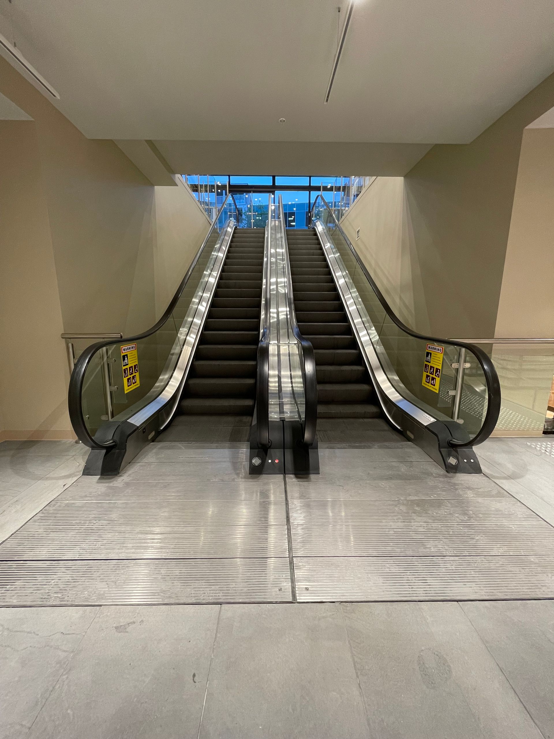 Two parallel escalators ascend, glass sides, metal steps, reaching an upper level with daylight.
