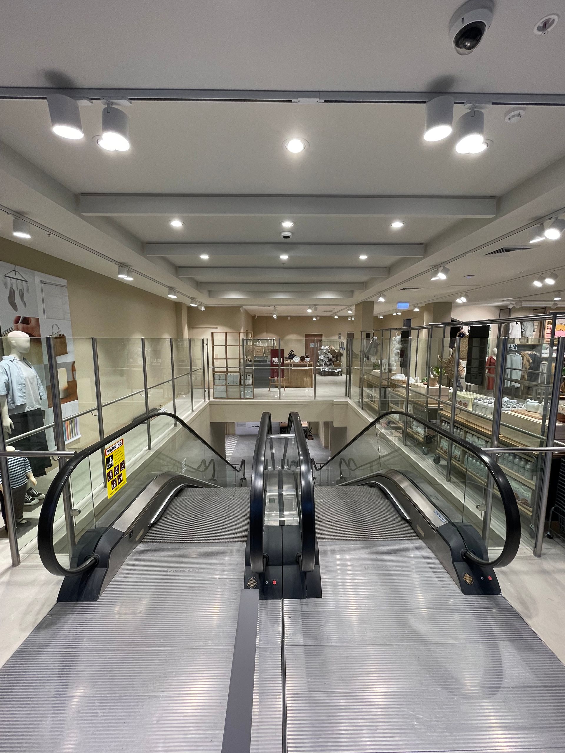 An empty escalator in a store, leading to a second level. Glass railings and overhead lighting.