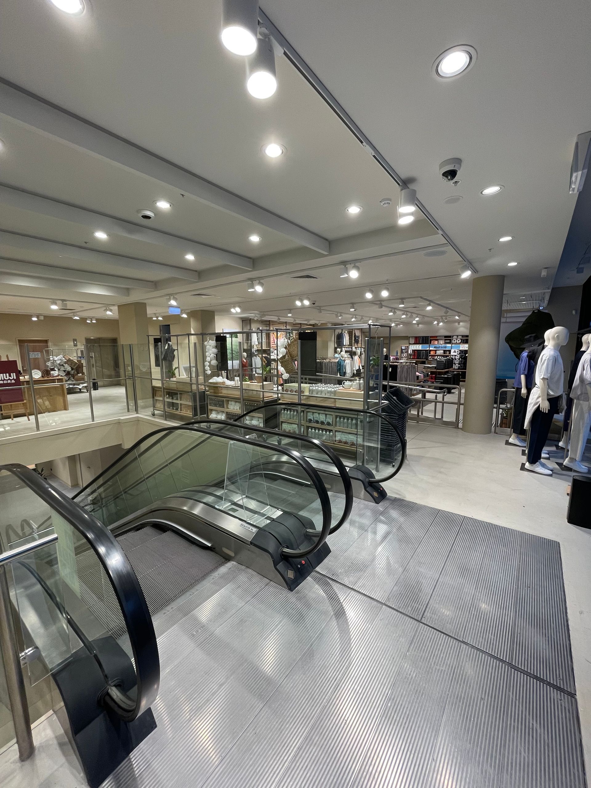 Escalator in a bright department store, leading to a display area with mannequins and merchandise.