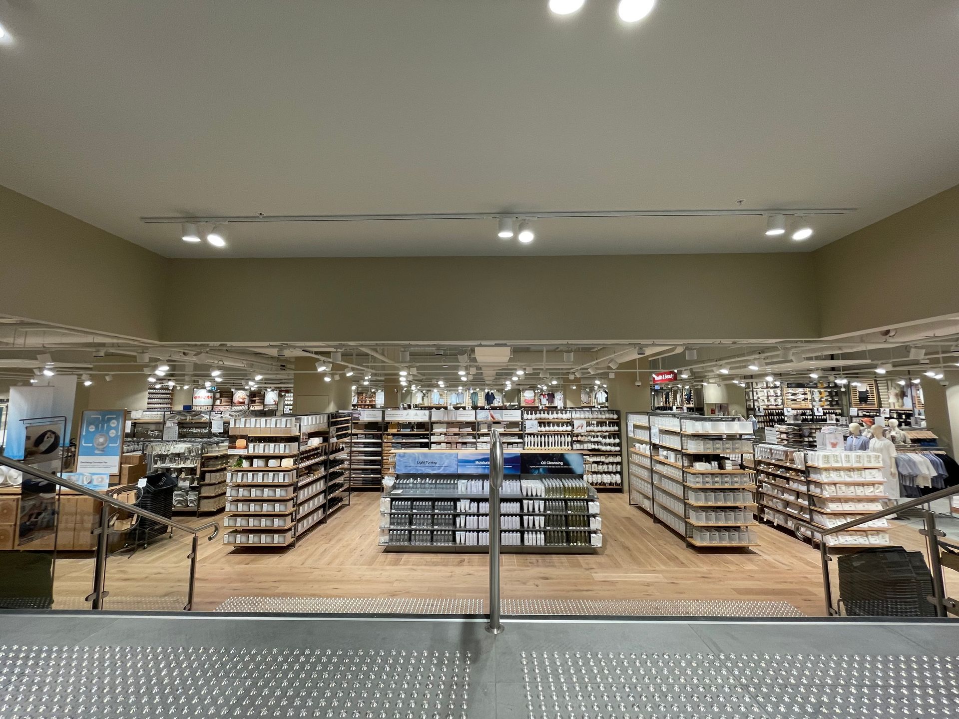 Interior of a well-lit store, featuring shelves of products, a wooden floor, and a metal handrail in the foreground.
